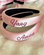 Personalized pink silk padded headbands with rhinestone names displayed on a soft plush background.