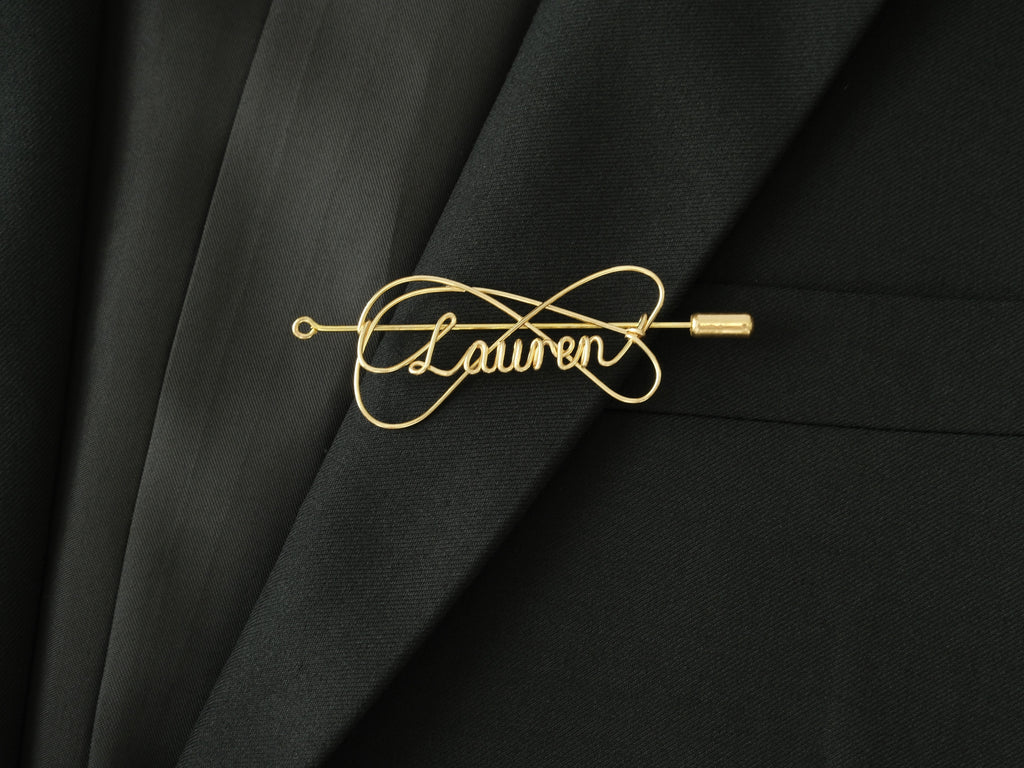 Custom name brooch with elegant double-line intertwined design, in gold-plated copper on a black jacket.