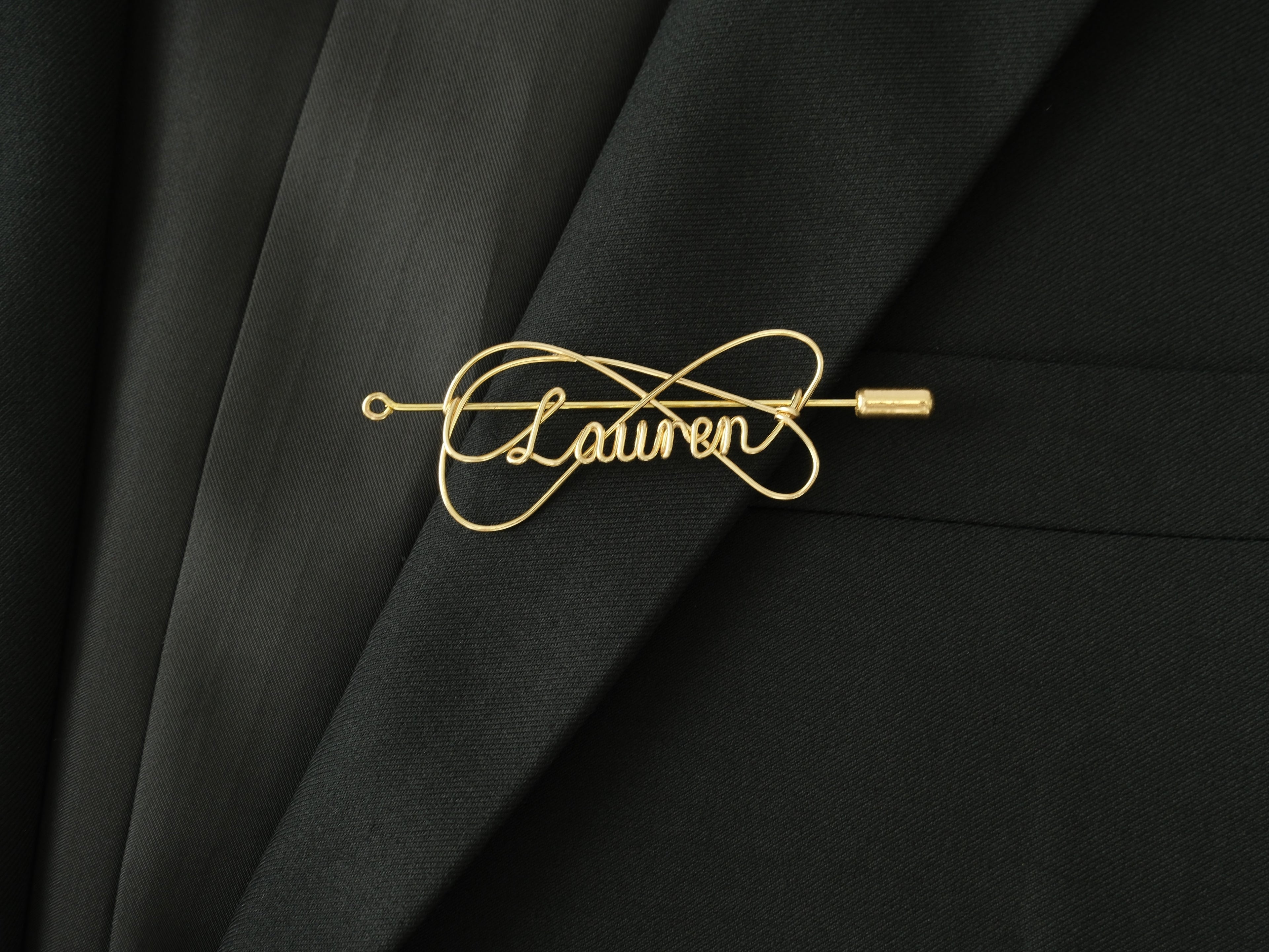 Custom name brooch with elegant double-line intertwined design, in gold-plated copper on a black jacket.