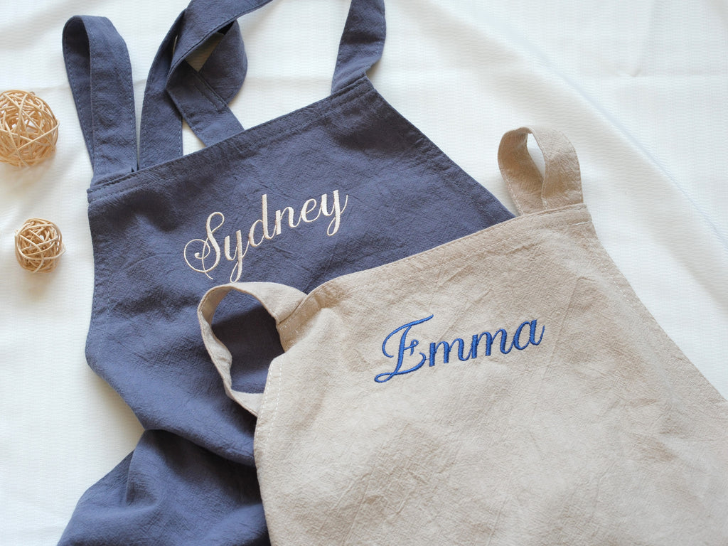 Two personalized linen aprons in Slate Blue and Oat Beige with "Sydney" and "Emma" embroidered in cursive.