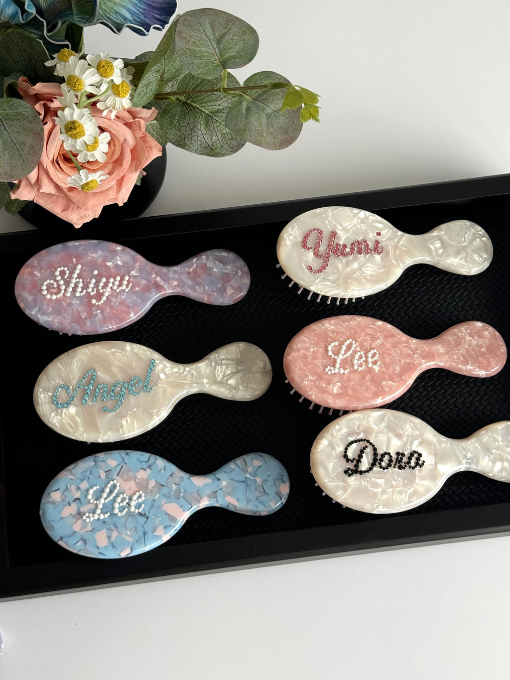 Collection of personalized mini hair brushes with glittering rhinestone names on marble-effect textures.