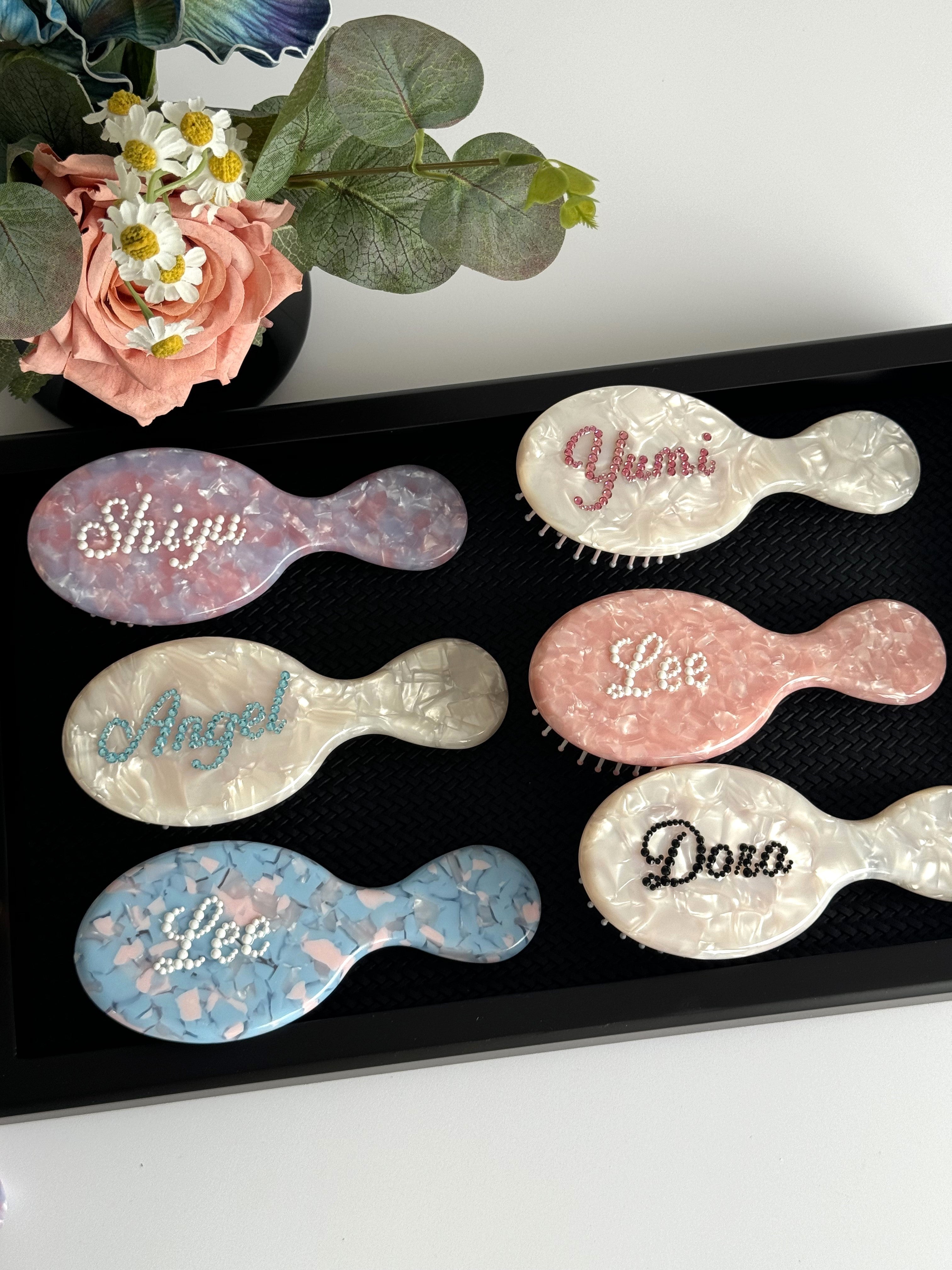 Collection of personalized mini hair brushes with glittering rhinestone names on marble-effect textures.