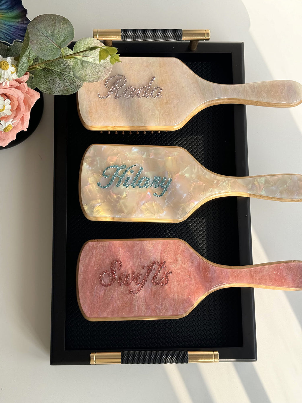 Personalized marble-effect paddle brushes with rhinestone names, captured under natural sunlight.