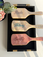 Personalized marble-effect paddle brushes with rhinestone names, captured under natural sunlight.