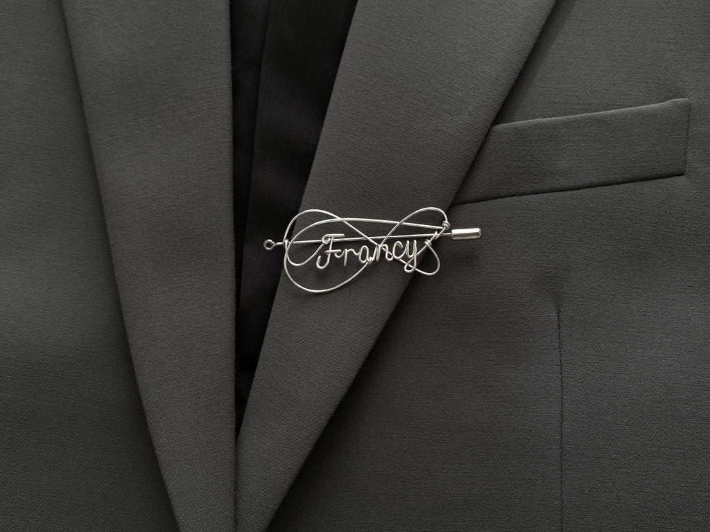 Personalized silver-plated name pin with an elegant double-intertwined line design on a dark green jacket.