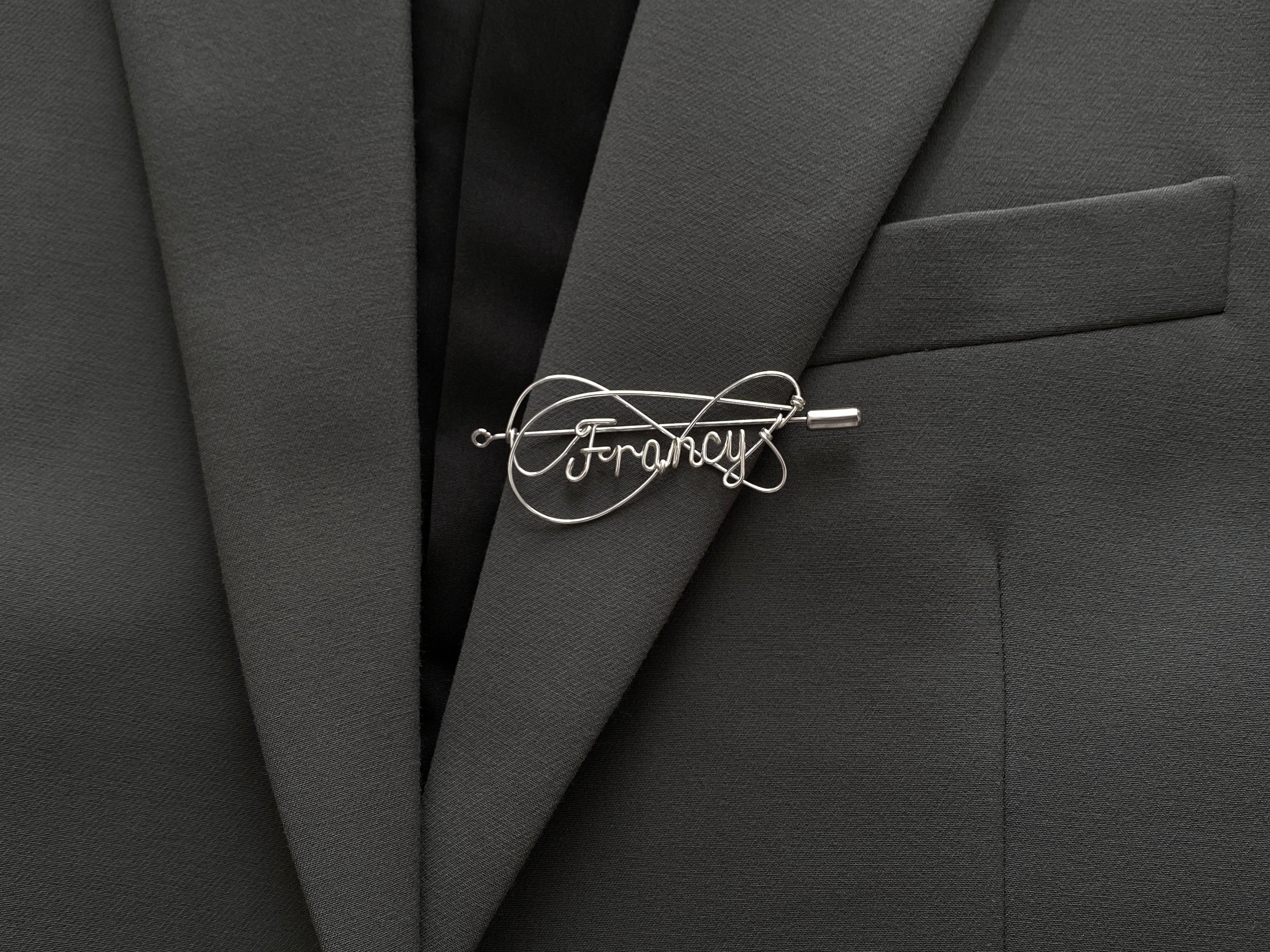 Personalized silver-plated name pin with an elegant double-intertwined line design on a dark green jacket.