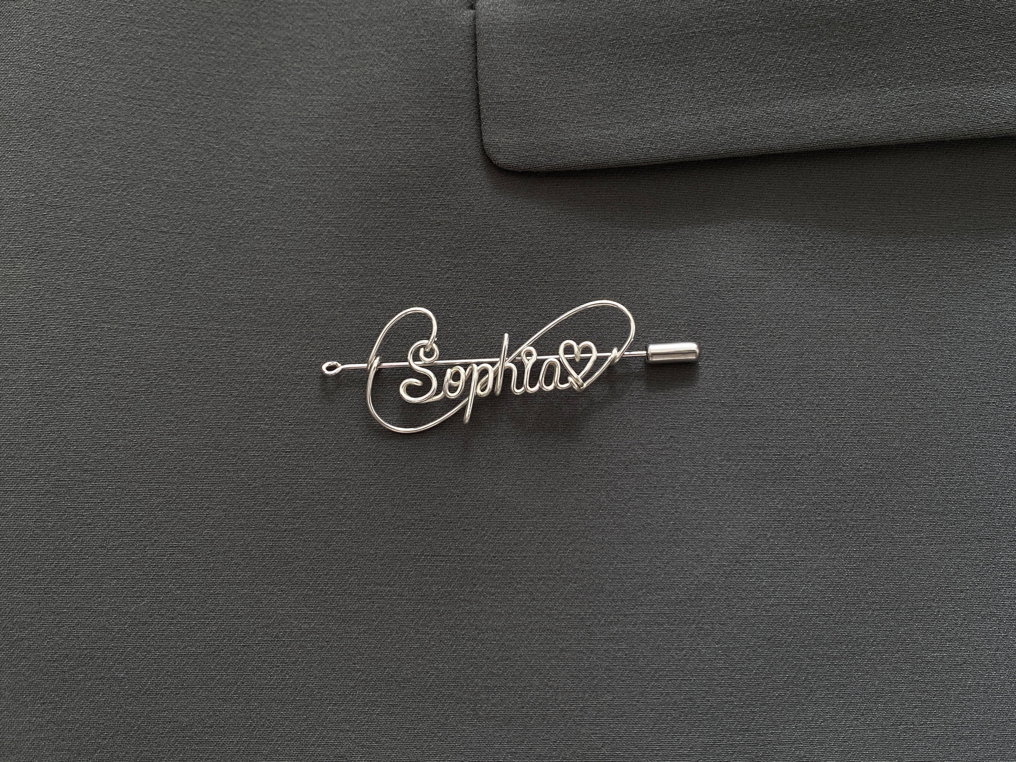 Personalized silver-plated name pin with a heart symbol on a dark green jacket, perfect gift idea.