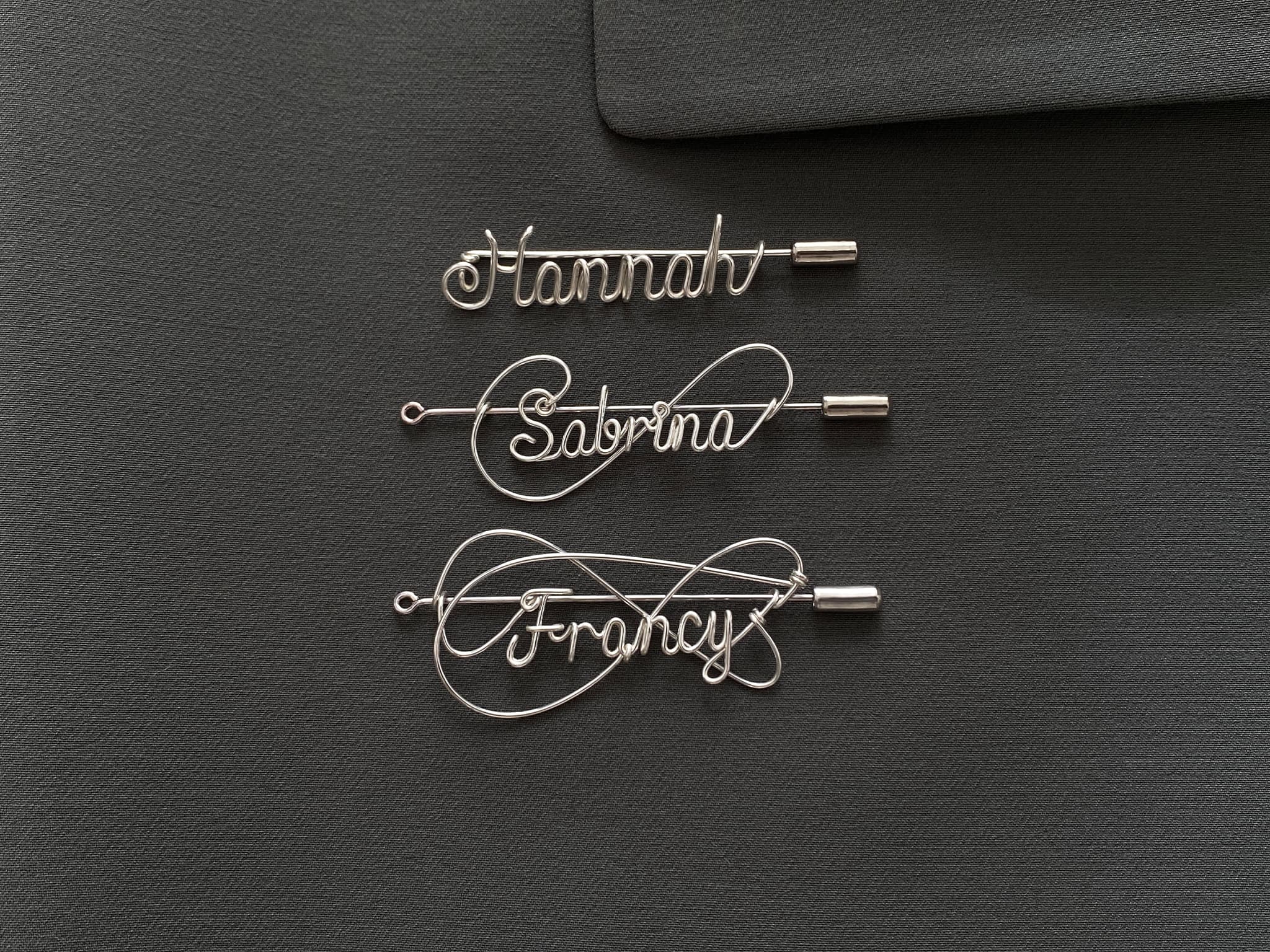 Three styles of personalized silver-plated name pins on a dark green jacket, handcrafted design.