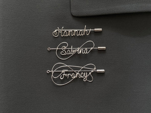 Three styles of personalized silver-plated name pins on a dark green jacket, handcrafted design.
