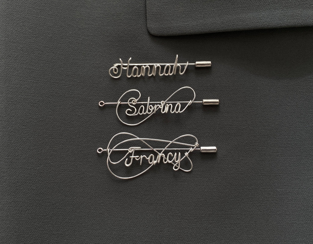Three styles of personalized silver-plated name pins on a dark green jacket, handcrafted design.