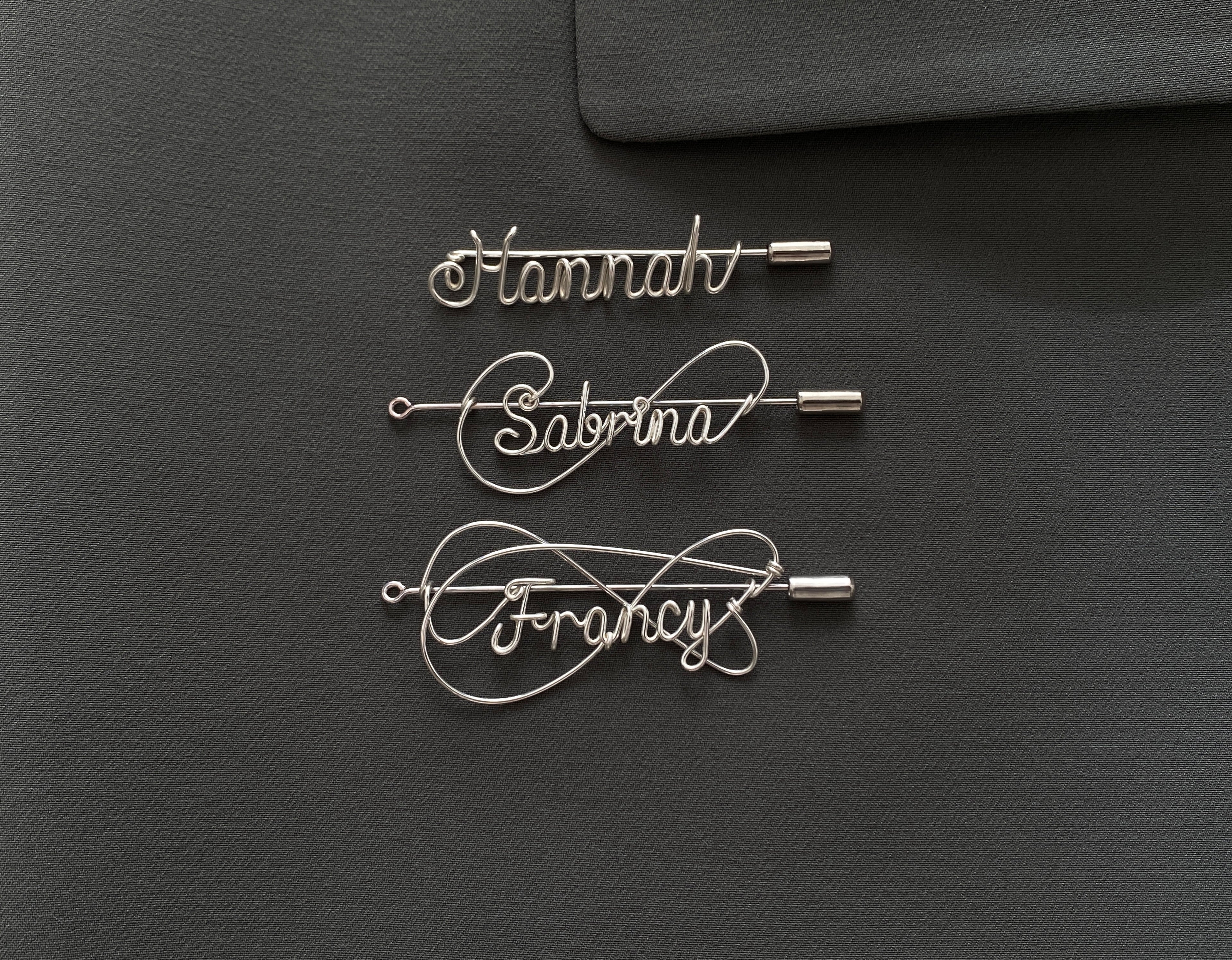 Three styles of personalized silver-plated name pins on a dark green jacket, handcrafted design.