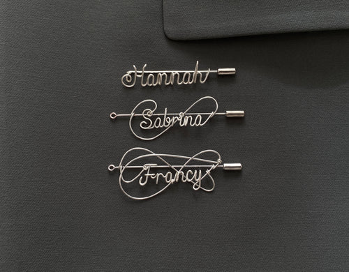 Three styles of personalized silver-plated name pins on a dark green jacket, handcrafted design.